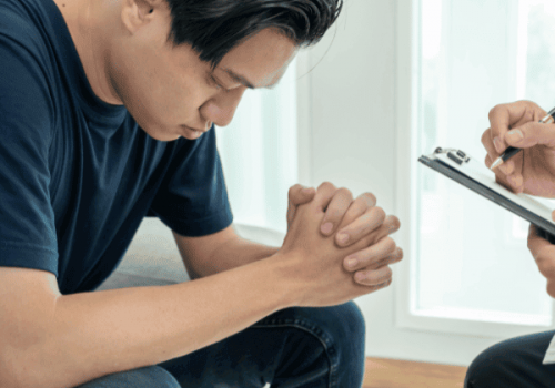 Young man in rehab treatment with his head down and hands folded while meeting with a therapist