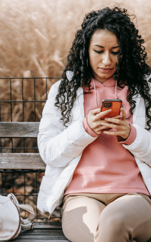 Woman sitting on a bench outdoors checking her phone for ComPsych addiction rehab coverage