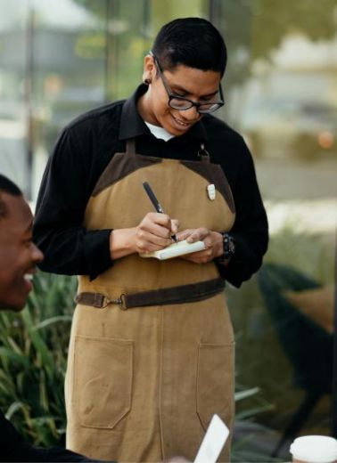 A smiling restaurant waiter is outdoors taking an order from two guests, one of whom is in partial view